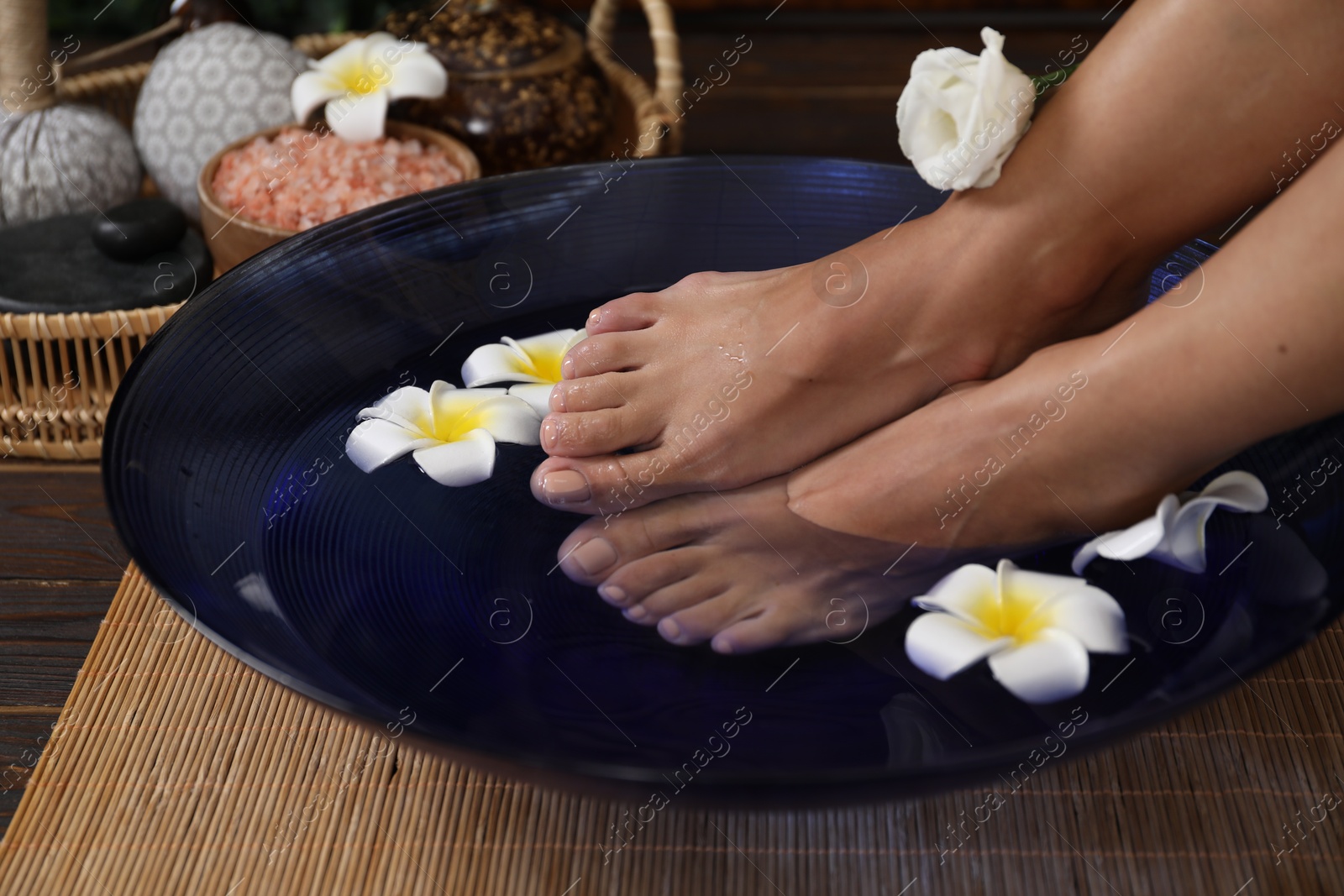 Photo of Woman soaking her feet in bowl with water and plumeria flowers on floor, closeup. Spa treatment