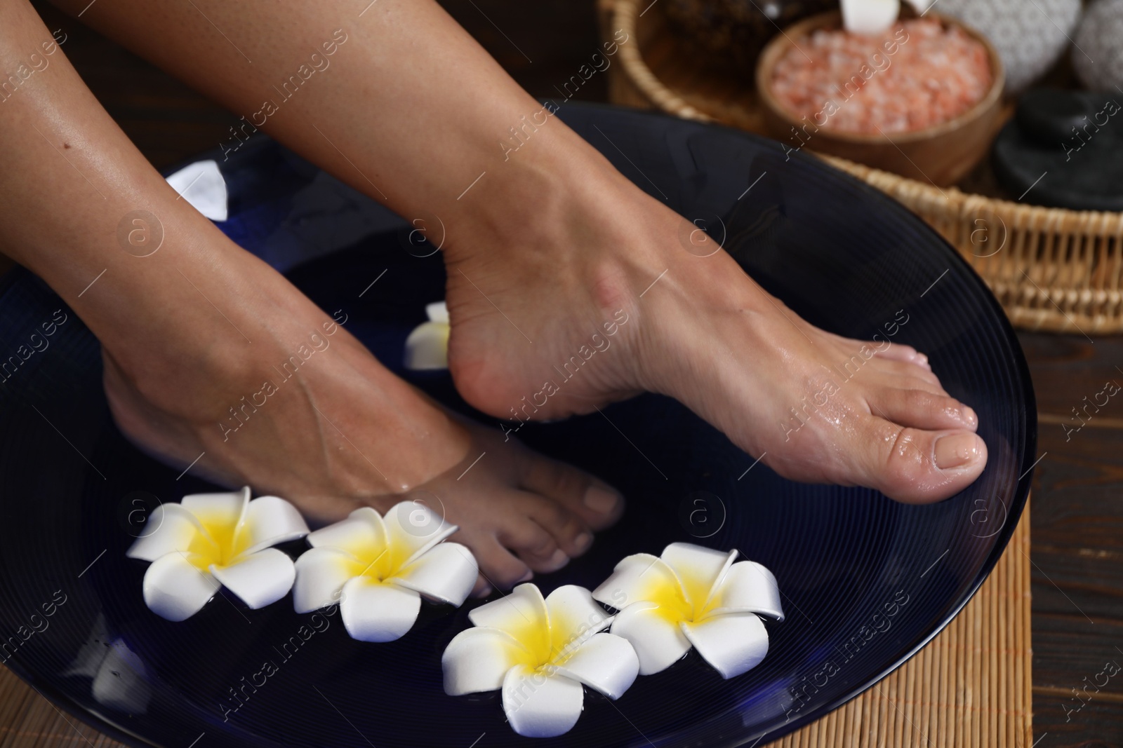 Photo of Woman soaking her feet in bowl with water and plumeria flowers on floor, closeup. Spa treatment