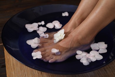 Photo of Woman soaking her feet in bowl with water and flowers on floor, closeup. Spa treatment