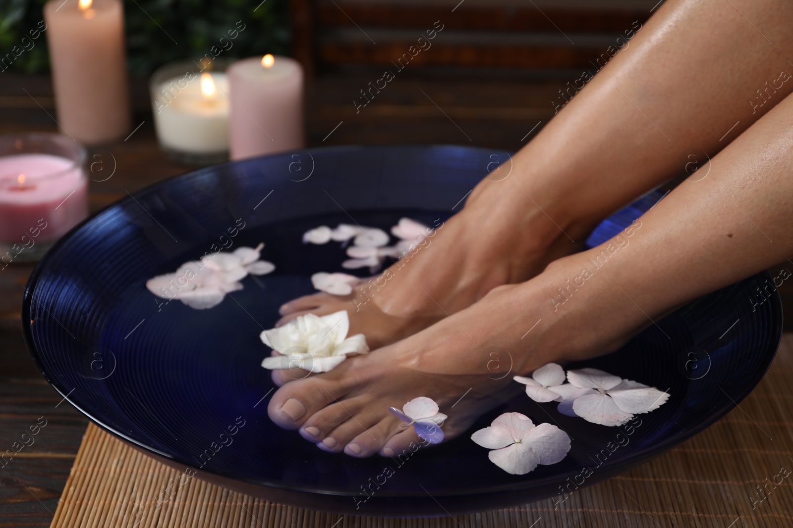 Photo of Woman soaking her feet in bowl with water and flowers on floor, closeup. Spa treatment