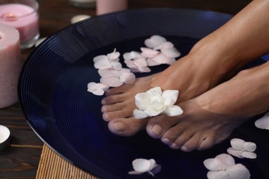 Photo of Woman soaking her feet in bowl with water and flowers on floor, closeup. Spa treatment