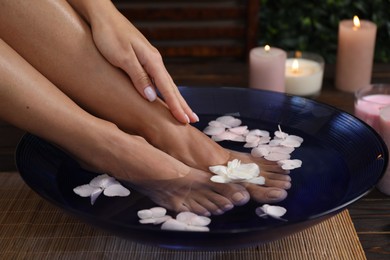 Photo of Woman soaking her feet in bowl with water and flowers on floor, closeup. Spa treatment