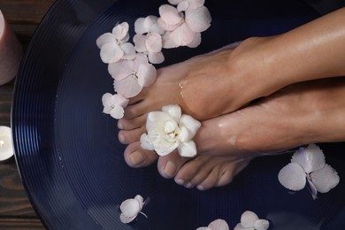 Photo of Woman soaking her feet in bowl with water and flowers on floor, top view. Spa treatment