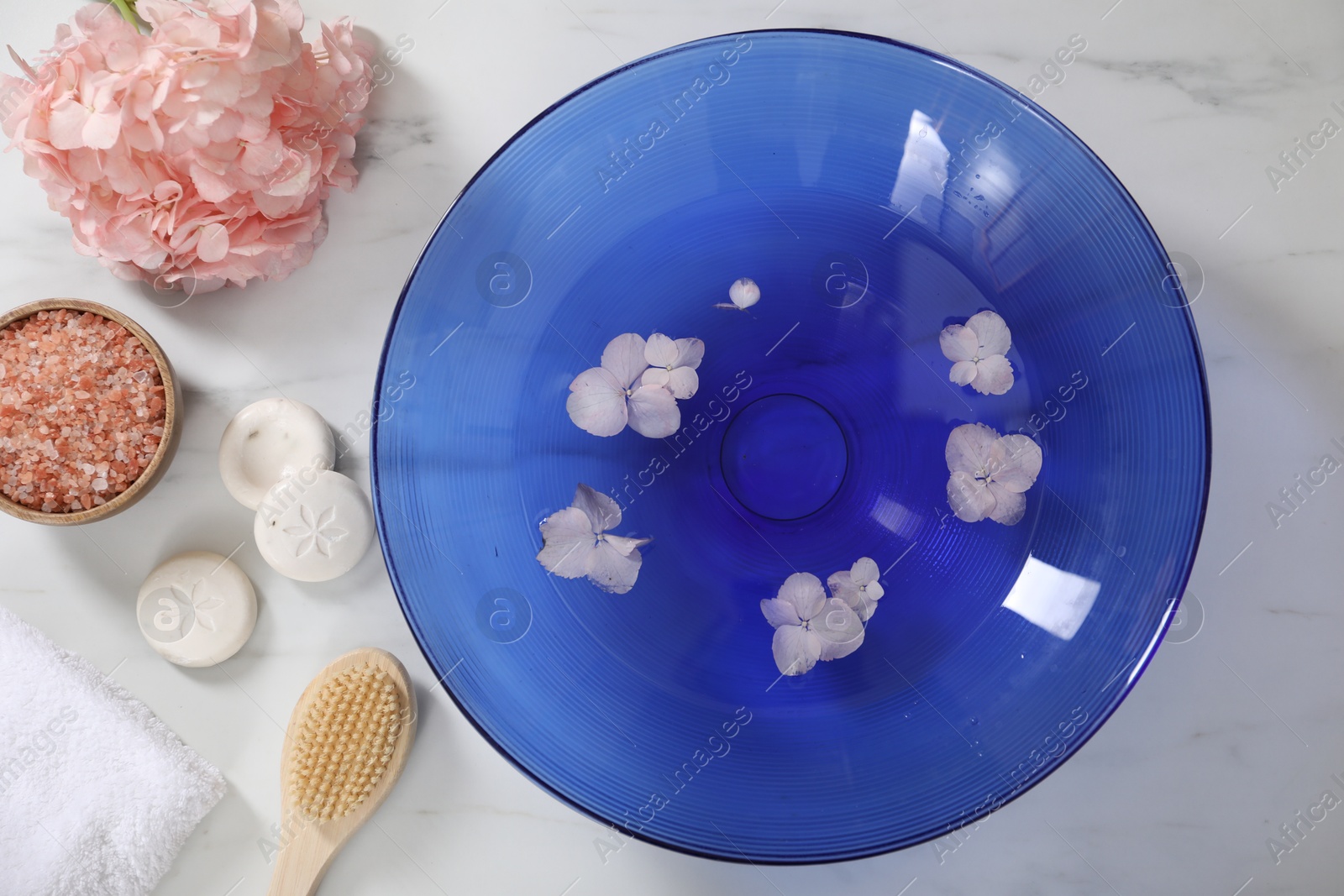 Photo of Bowl of water with flowers and different spa supplies on white marble table, flat lay