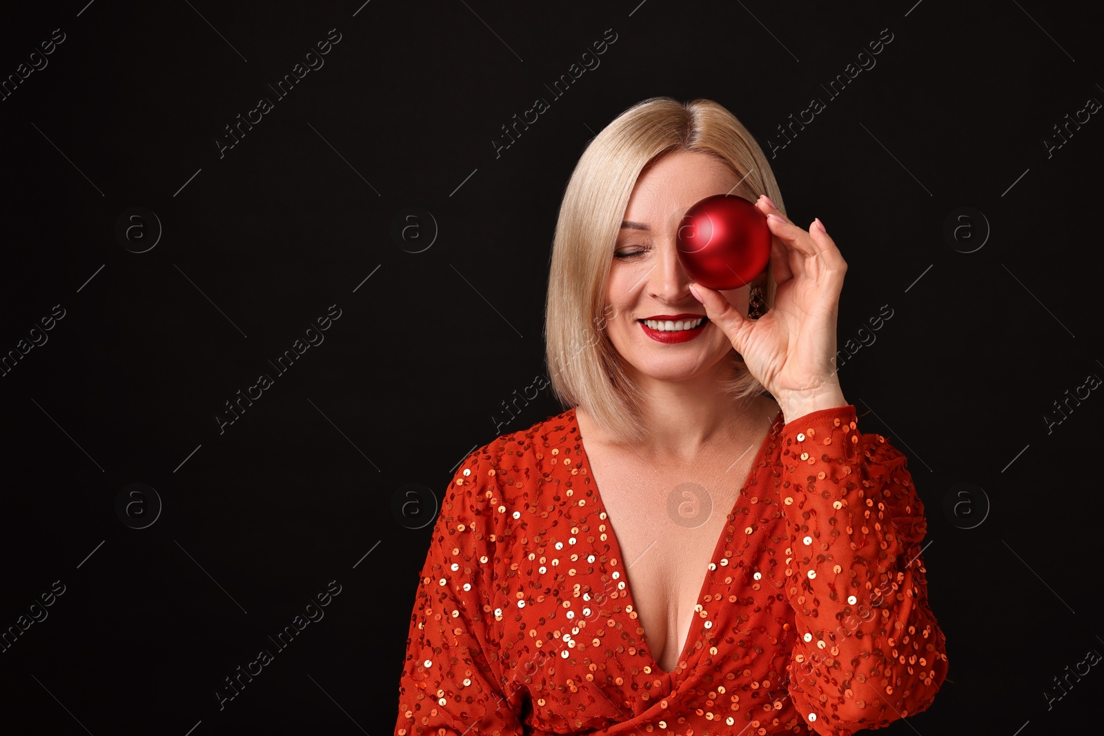 Photo of Smiling woman with perfect makeup holding Christmas bauble on black background. Space for text