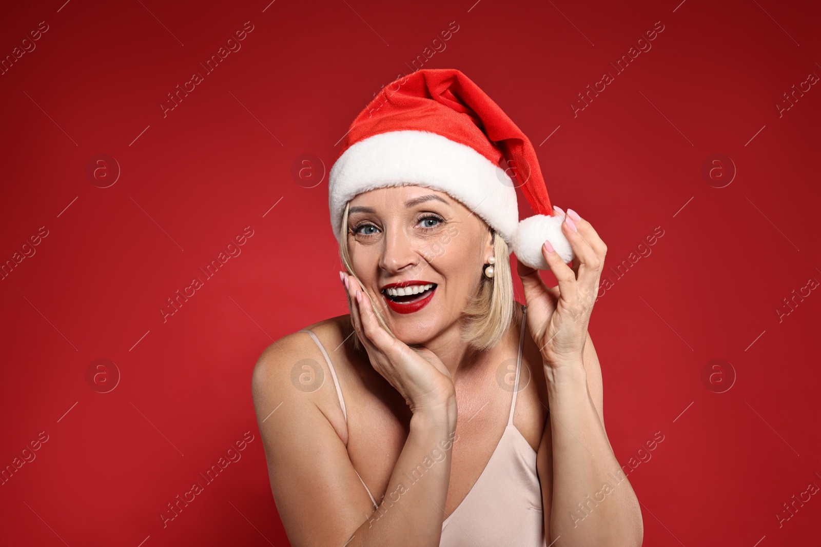 Photo of Smiling woman with perfect makeup in Santa hat celebrating Christmas on red background