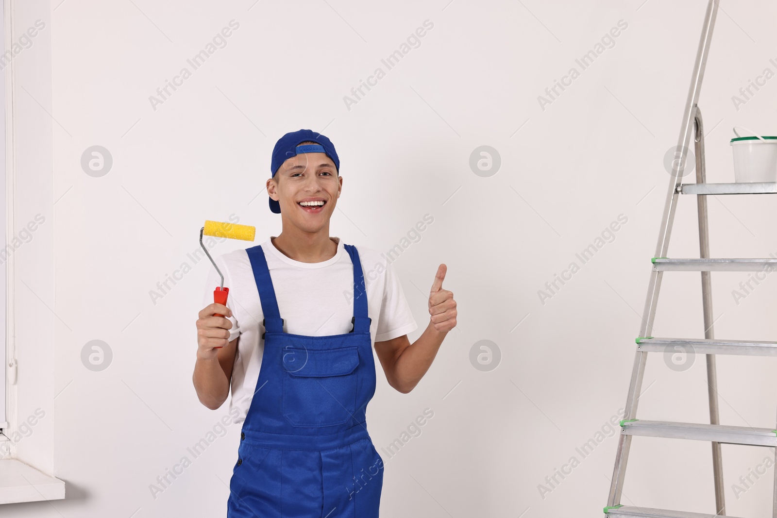 Photo of Smiling handyman with paint roller showing thumbs up indoors