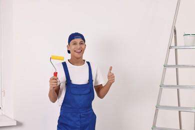 Photo of Smiling handyman with paint roller showing thumbs up indoors