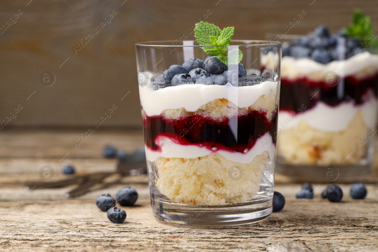 Photo of Tasty trifle dessert. Sponge cake, blueberries, jam and whipped cream in glasses on wooden table, closeup