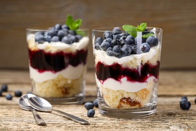 Photo of Tasty trifle dessert. Sponge cake, blueberries, jam and whipped cream in glasses on wooden table, closeup