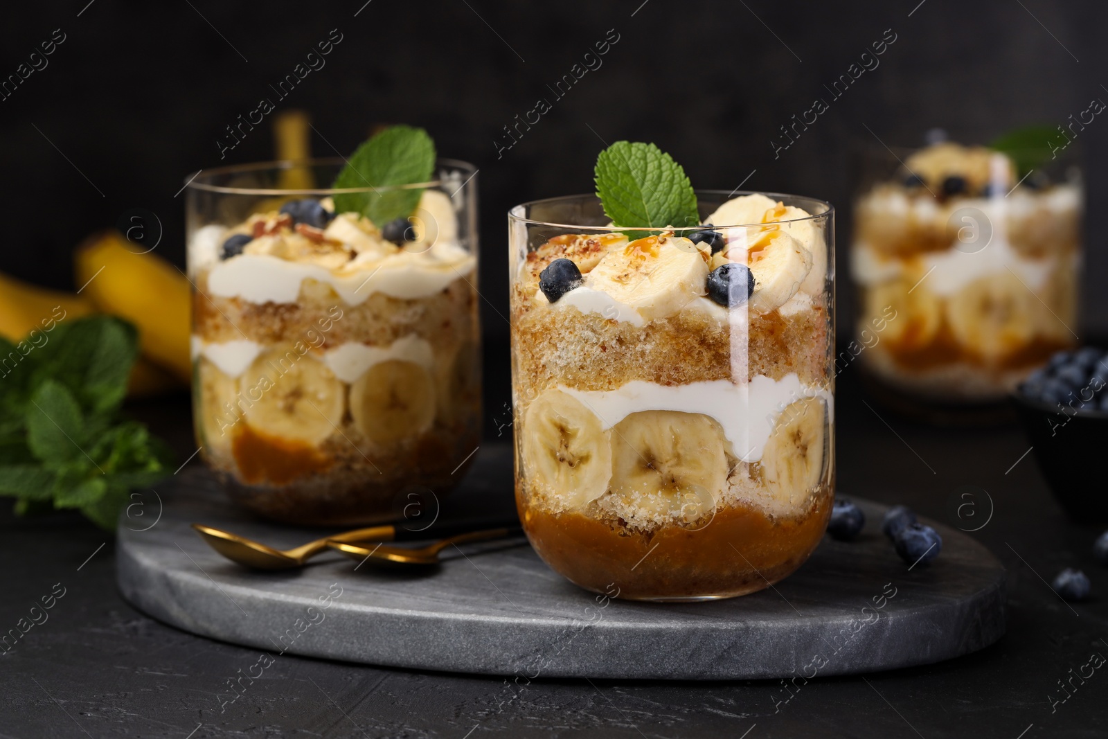 Photo of Tasty trifle dessert. Sponge cake, blueberries, banana and whipped cream in glasses on black table