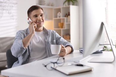 Happy man talking by smartphone at desk with computer indoors. Home office