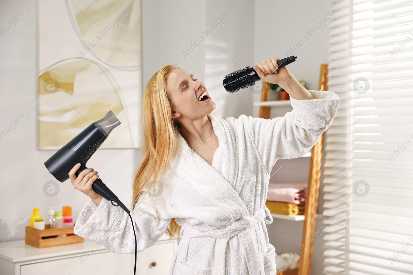 Photo of Beautiful young woman drying her hair and singing into brush in bathroom