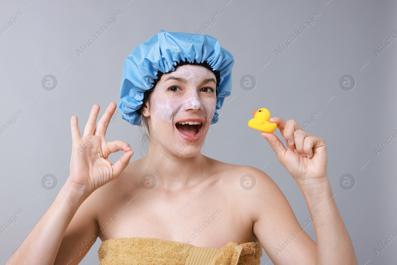 Photo of Woman with shower cap and bath duck showing ok gesture on grey background