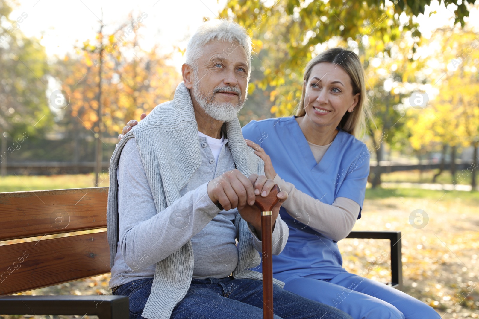 Photo of Caregiver assisting senior man on wooden bench in park. Home health care service