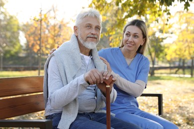 Photo of Caregiver assisting senior man on wooden bench in park. Home health care service