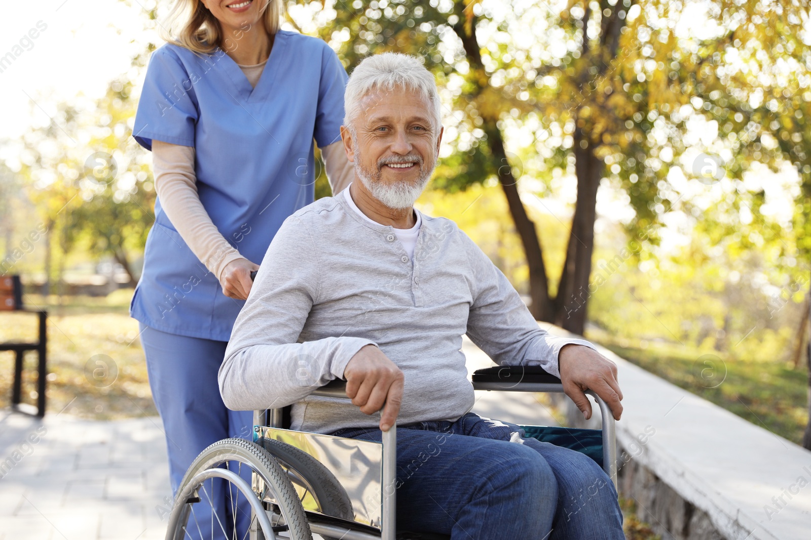 Photo of Caregiver assisting senior man on wheelchair in park. Home health care service