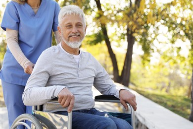 Photo of Caregiver assisting senior man on wheelchair in park. Home health care service