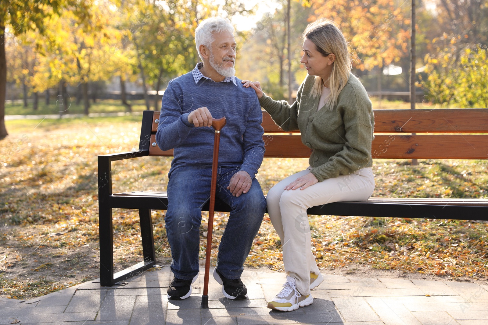 Photo of Caregiver assisting senior man on wooden bench in park. Home health care service