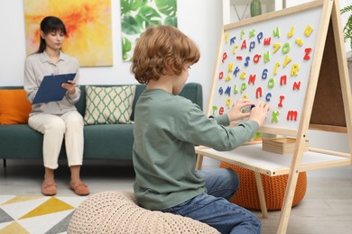 Photo of Boy assembling letters on magnetic board while psychologist taking notes indoors, selective focus