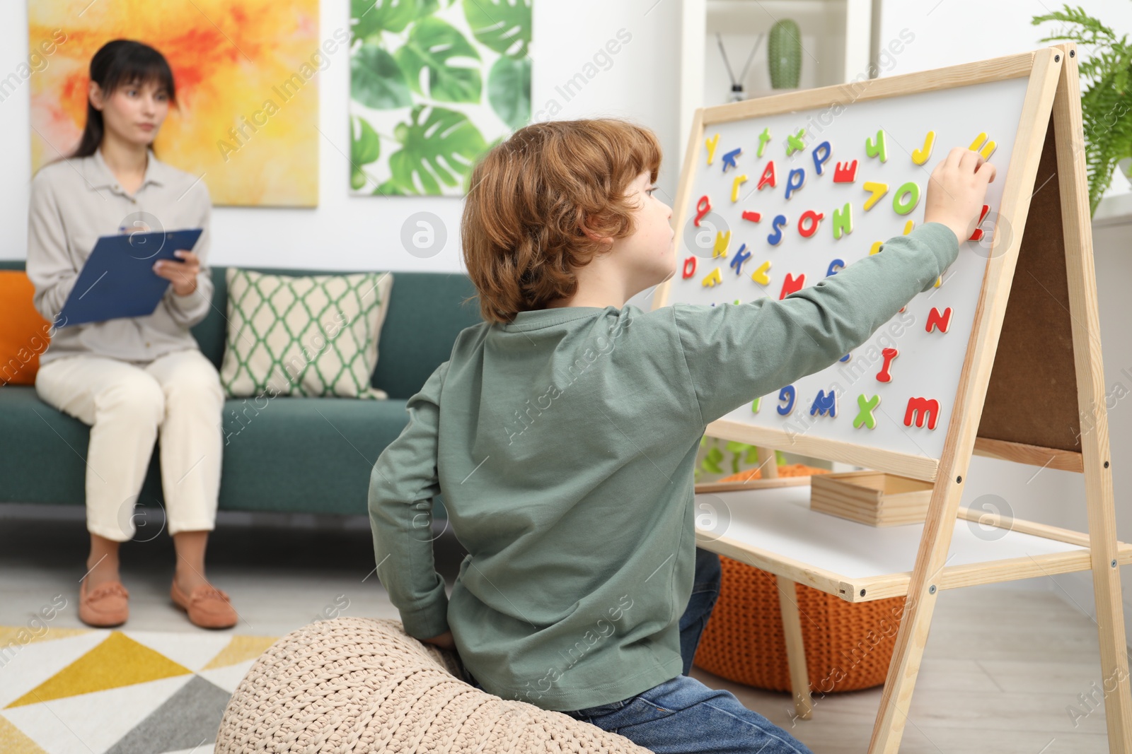 Photo of Boy assembling letters on magnetic board while psychologist taking notes indoors, selective focus