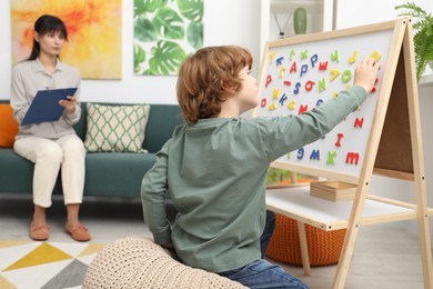 Boy assembling letters on magnetic board while psychologist taking notes indoors, selective focus