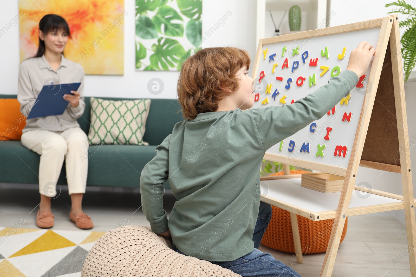 Photo of Boy assembling letters on magnetic board while psychologist taking notes indoors, selective focus