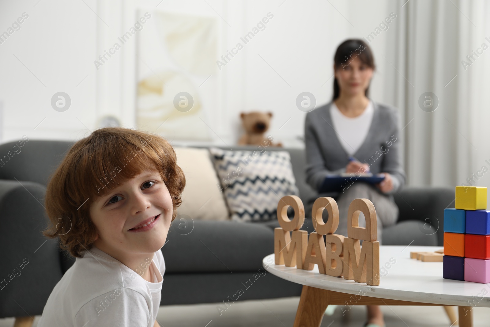 Photo of Boy playing with wooden letters at table while psychologist taking notes, selective focus