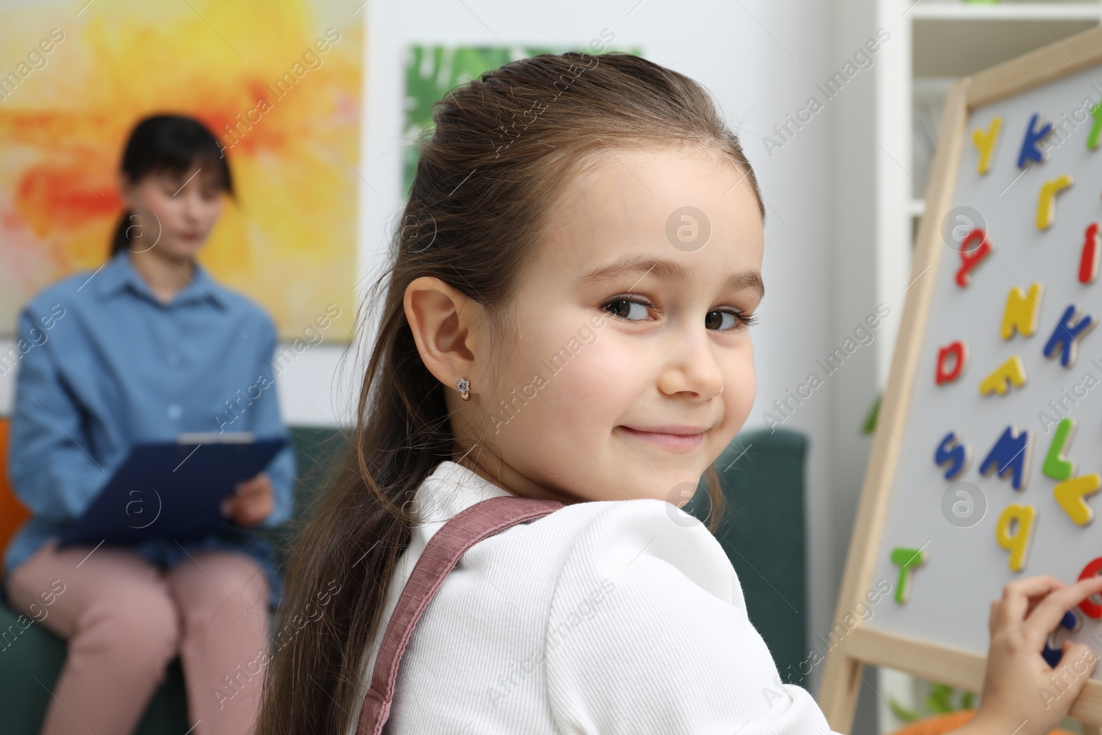 Photo of Girl assembling letters on magnetic board while psychologist taking notes indoors, selective focus