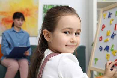 Photo of Girl assembling letters on magnetic board while psychologist taking notes indoors, selective focus