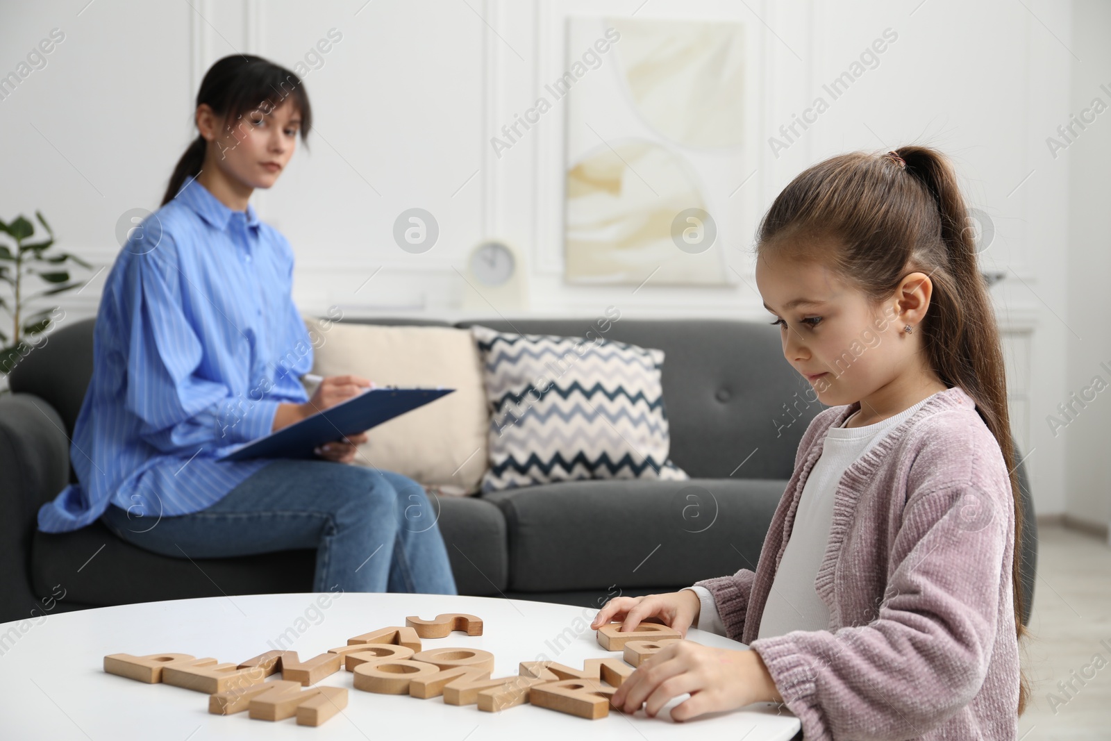 Photo of Girl playing with letters at table while psychologist taking notes, selective focus