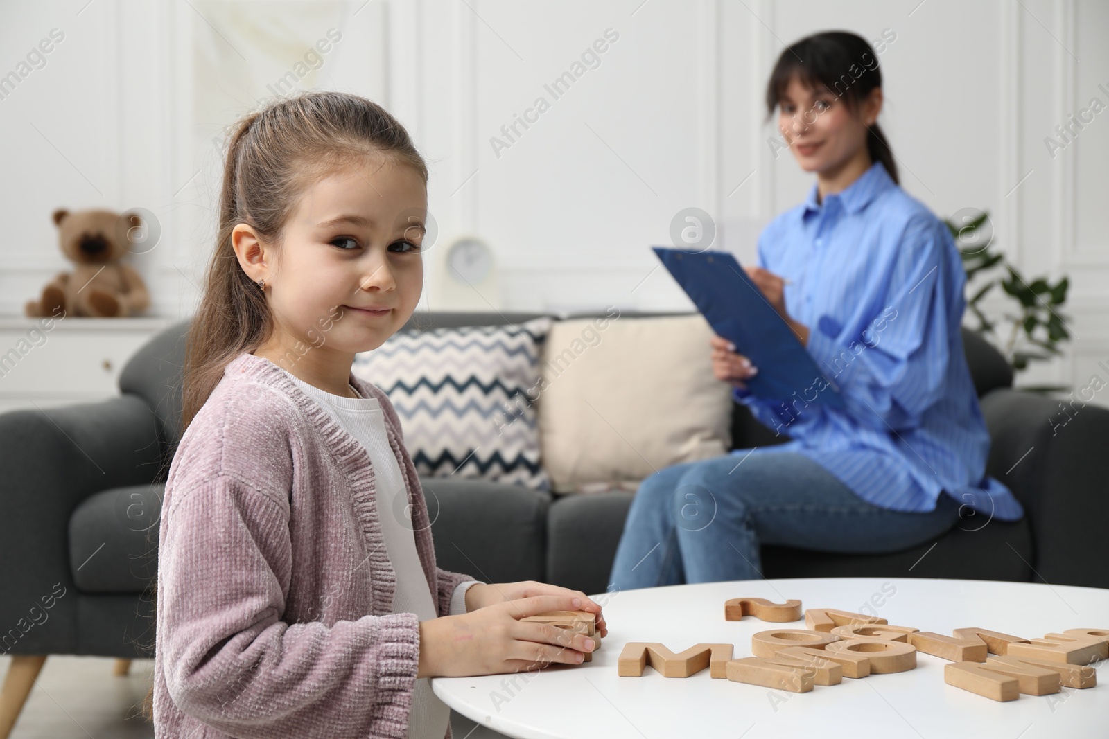 Photo of Girl playing with letters at table while psychologist taking notes, selective focus