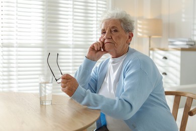 Photo of Sad senior woman feeling lonely at table with glass of water indoors