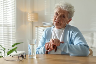 Photo of Thoughtful senior woman feeling lonely at table with glass of water indoors