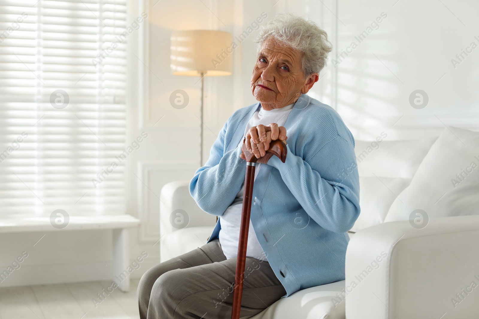 Photo of Lonely senior woman with walking cane sitting on sofa indoors. Space for text