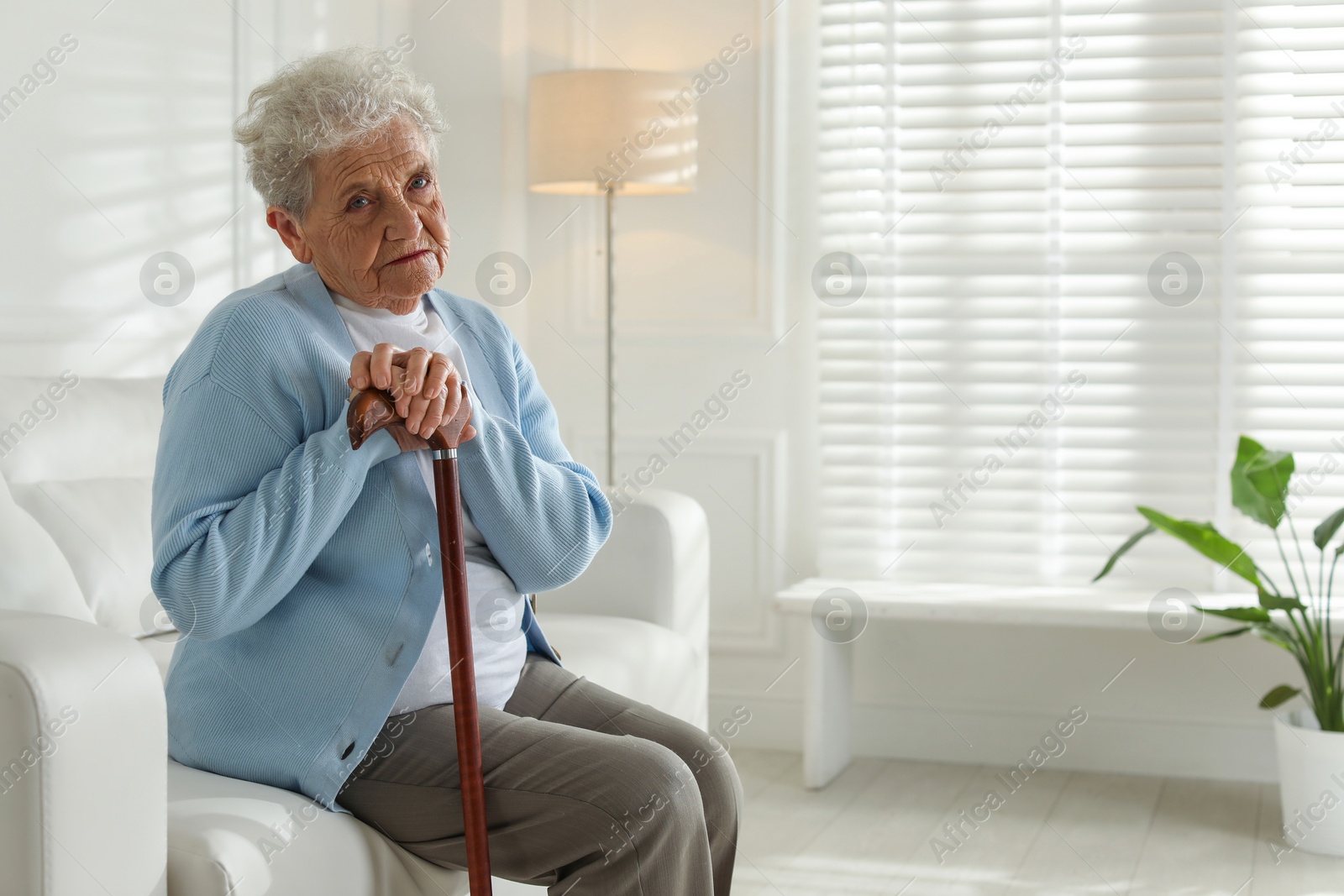 Photo of Lonely senior woman with walking cane sitting on sofa indoors. Space for text