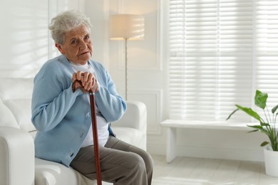 Photo of Lonely senior woman with walking cane sitting on sofa indoors. Space for text