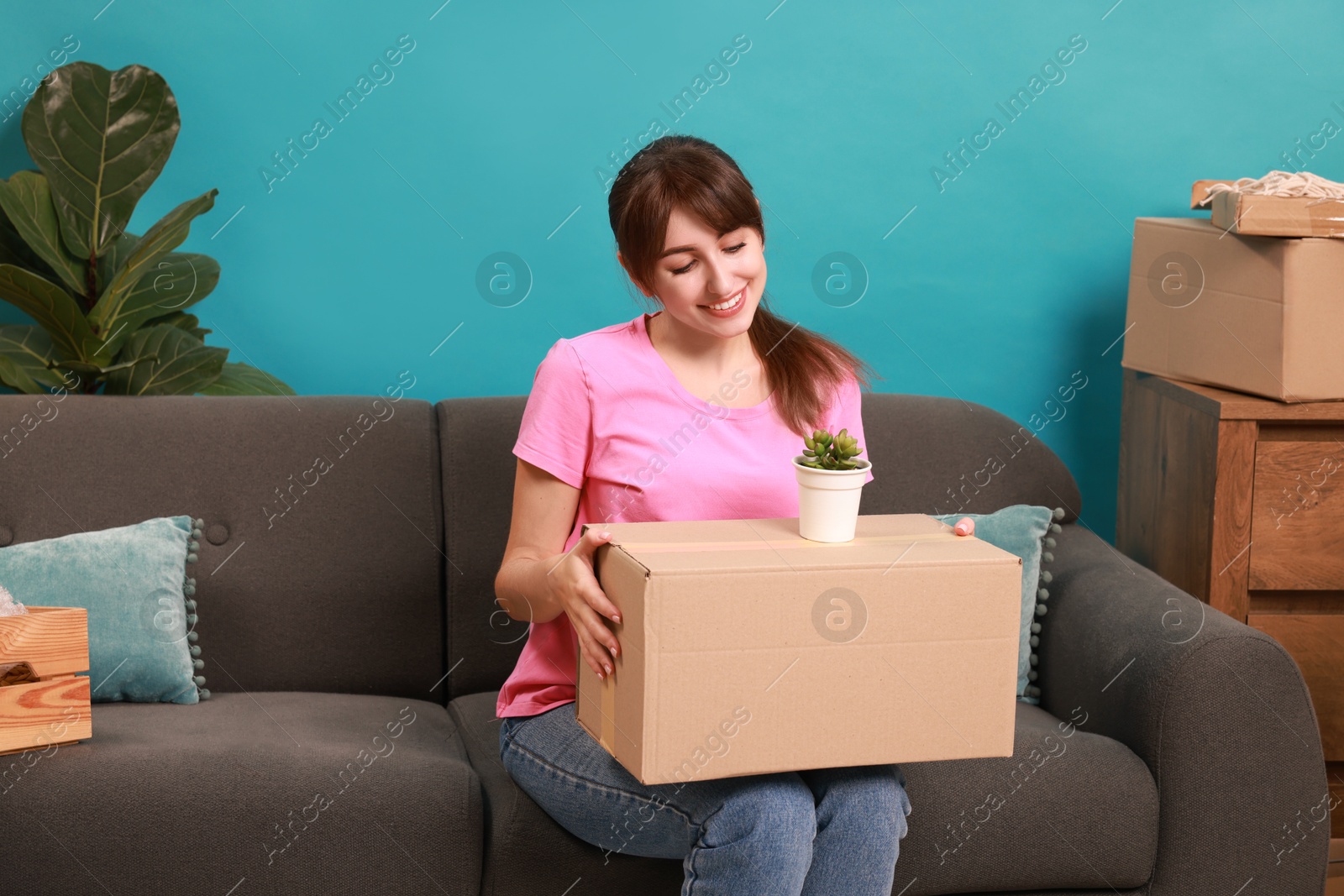 Photo of Happy woman with moving box and houseplant on sofa in new apartment. Housewarming party