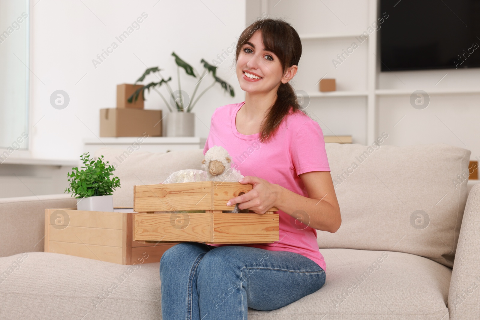 Photo of Happy woman holding wooden crate with stuff on sofa in new apartment. Housewarming party