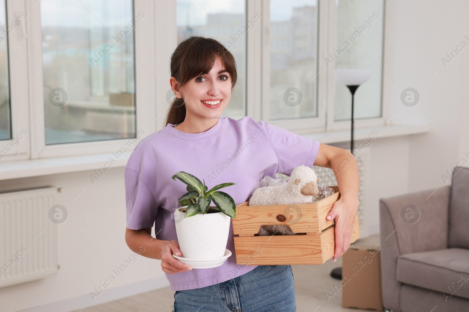 Photo of Happy woman holding wooden crate with stuff and houseplant in new apartment. Housewarming party