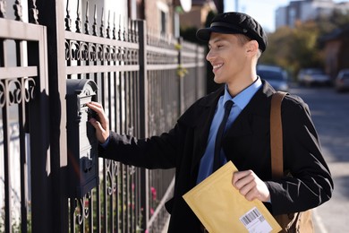 Happy postman putting envelope into mail box outdoors