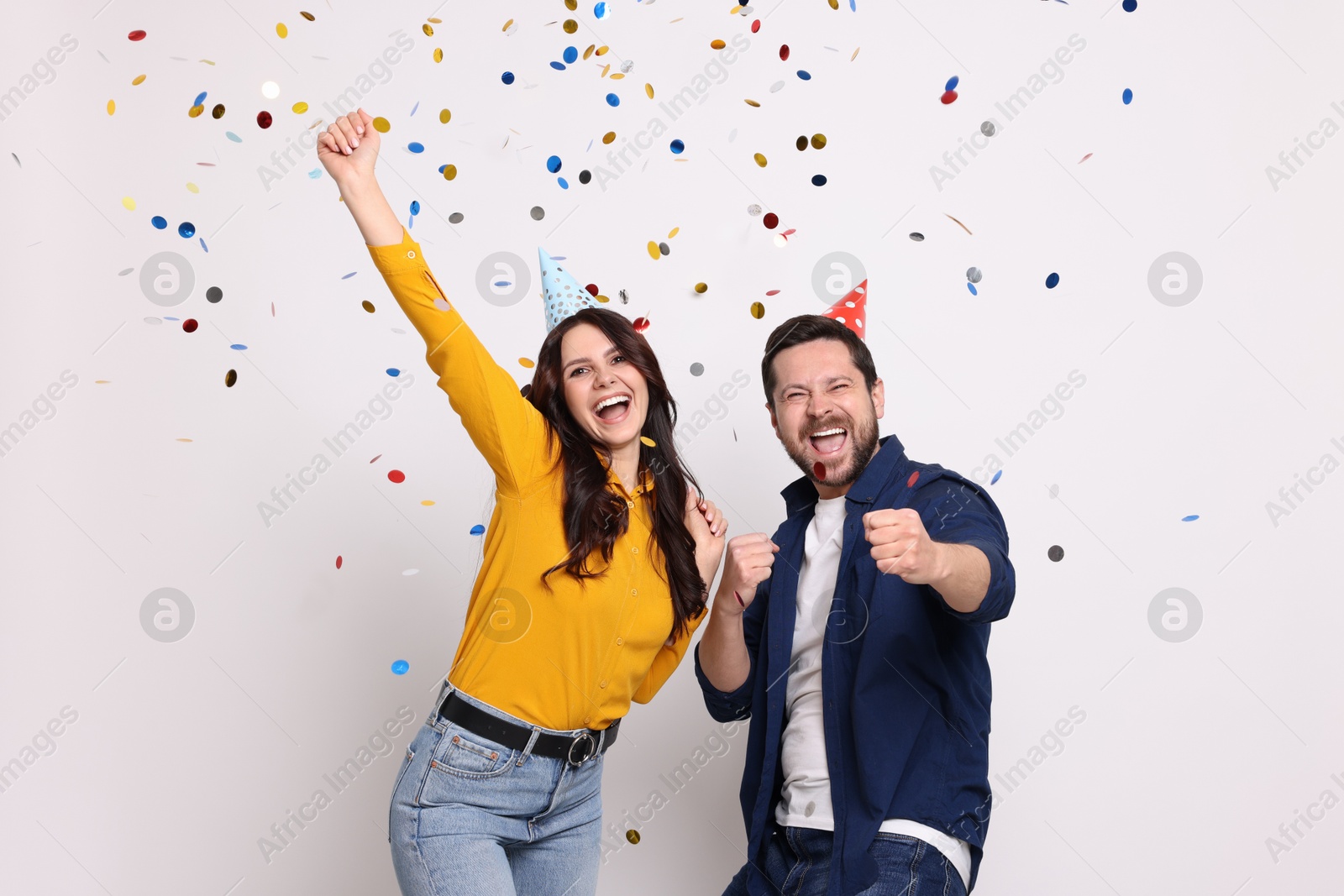 Photo of Happy friends in conical paper hats having fun under falling confetti on white background. Surprise party