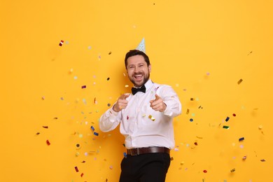 Happy man in conical paper hat and flying confetti on orange background. Surprise party