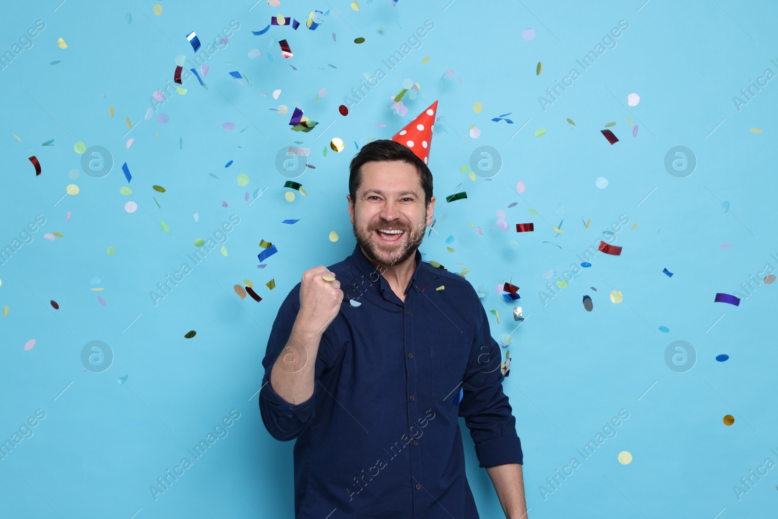 Photo of Happy man in conical paper hat under falling confetti on light blue background. Surprise party