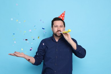 Photo of Man in conical paper hat with blower and flying confetti on light blue background. Surprise party