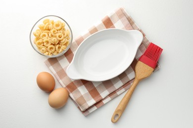 Photo of Ceramic casserole, raw pasta, eggs and brush on white table, flat lay