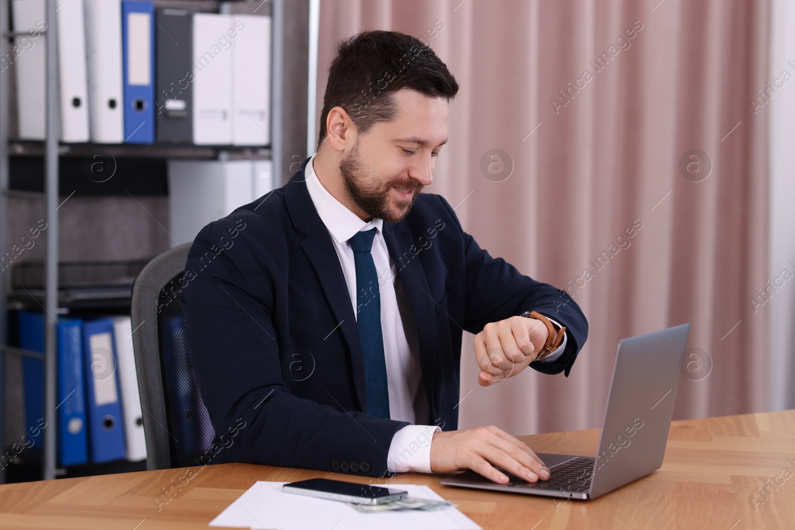 Photo of Banker checking time while working with laptop at wooden table in office