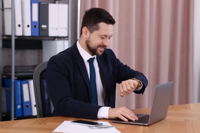 Photo of Banker checking time while working with laptop at wooden table in office