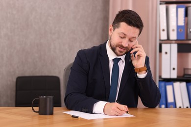 Photo of Banker talking on smartphone while working with documents at wooden table in office