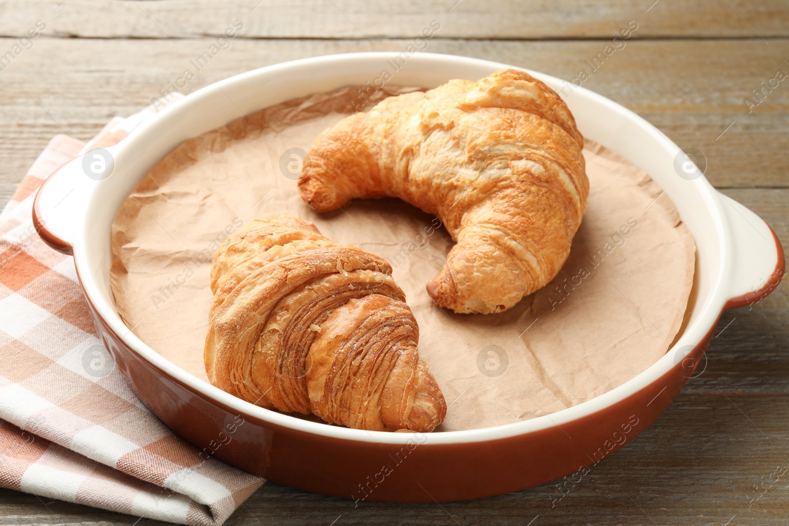 Photo of Ceramic casserole with croissants and baking parchment paper on wooden table, closeup